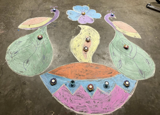 Colorful rangoli depicting two peacocks branching out from either side of a lit candle (diya).
