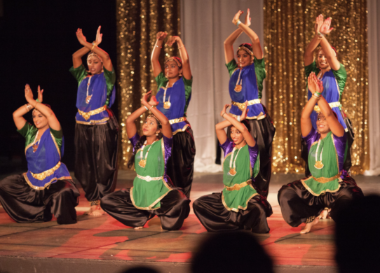 8 women classical dancers dressed in green and purple traditional tops and black pants strike a dance pose onstage.