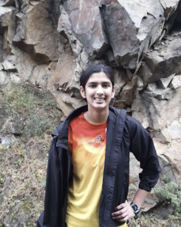 Pavni Jagpal smiling, wearing a red and yellow shirt and a black jacket, against the backdrop of a cliffside.