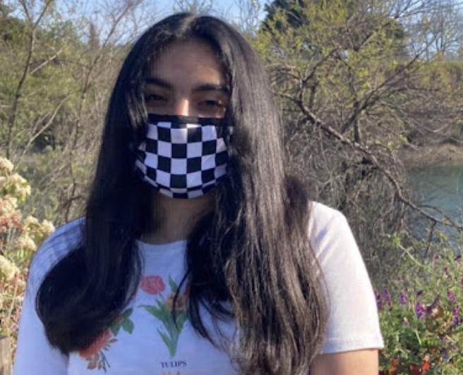 Hanya Hussain in a checkered mask and white shirt with a flower pattern against the backdrop of greenery and a lake.