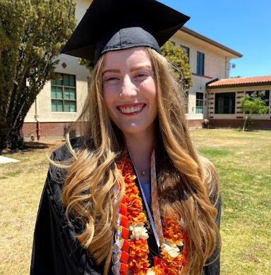 Ashley Tosh, wearing a black graduation cap, with a lei, medals, and a graduation gown, all against the backdrop of trees and Washington High School.