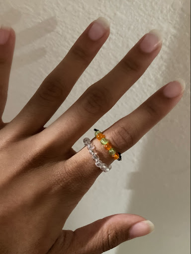 A hand, two beaded rings on the pointer finger—the bottom ring is made of clear beads and the top one alternates with green and orange beads, against the backdrop of a white wall.