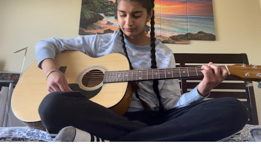 Sneha Kamboj, sitting criss cross applesauce, wearing a long-sleeved gray shirt and black leggings, playing her guitar in her room, against the backdrop of a yellow wall with a painting of a sunset.