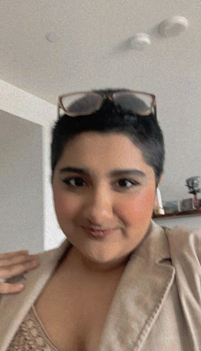Kavya Jolly in a cream-colored jacket and shirt, with brown-rimmed sunglasses, and an airpod.