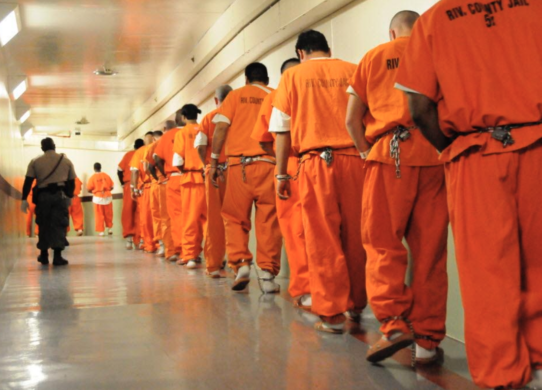 Prison inmates in orange jumpsuits in a line.